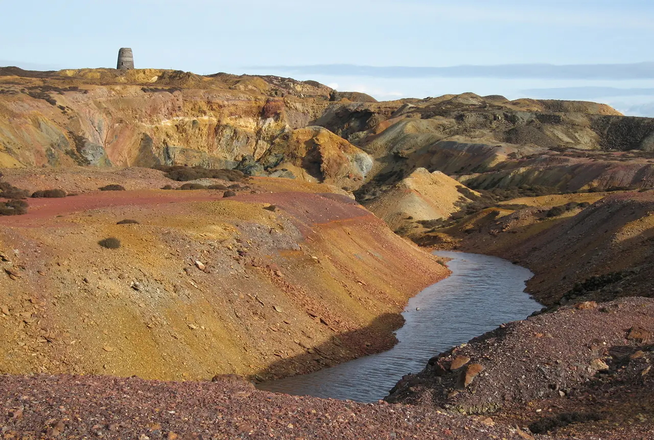 Hills of red, brown and yellow soil with water in the bottom of the valleys. A small tower is on the horizon.