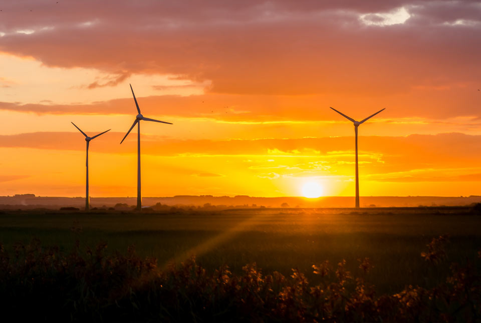 Tall wind turbines are silhouetted against an orange sky, with the sun setting on the horizon