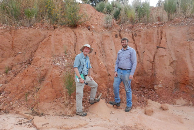 Two white men are standing in front of a low, sandy cliff
