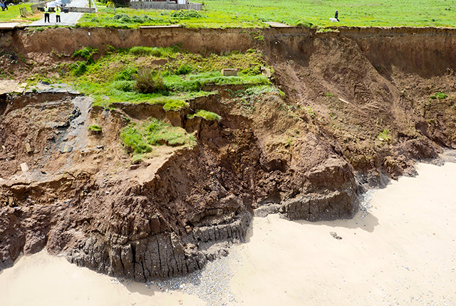 Landslide debris, which has fallen from the top of a cliff covered in grass to a sandy beach below. A broken tarmac road can be seen to the left, with two people in high visibility jackets looking down towards the rubble
