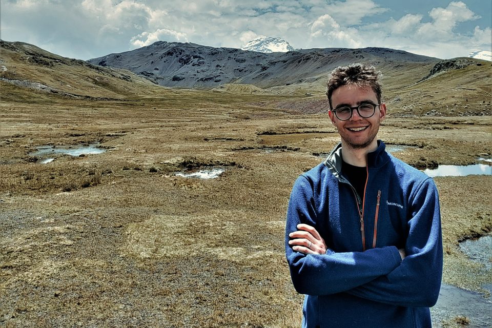 A white man in a blue jacket standing in front of a grassy plain with snowy mountains in the background. He is smiling broadly and has his arms folded.