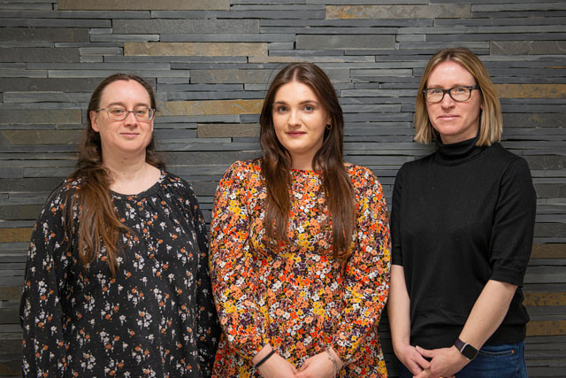 Three white women standing in front of a grey slate wall. Nicola on the left is wearing a black top with white flowers; she has long brown hair and glasses. Leah in the middle has long dark hair and is wearing an orange floral top. Angela on the right has bobbed blonde hair and is wearing glasses and a black jumper.