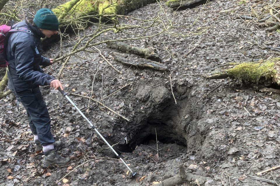 A person wearing a green hat and a blue coat standing on ground covered in leaves in front of a deep hole.