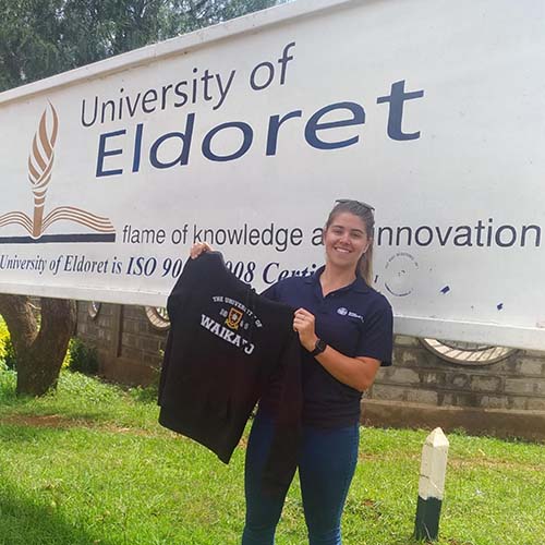 A woman stands in front of a sign for the University of Eldoret holding up a t shirt that says University of Waikato